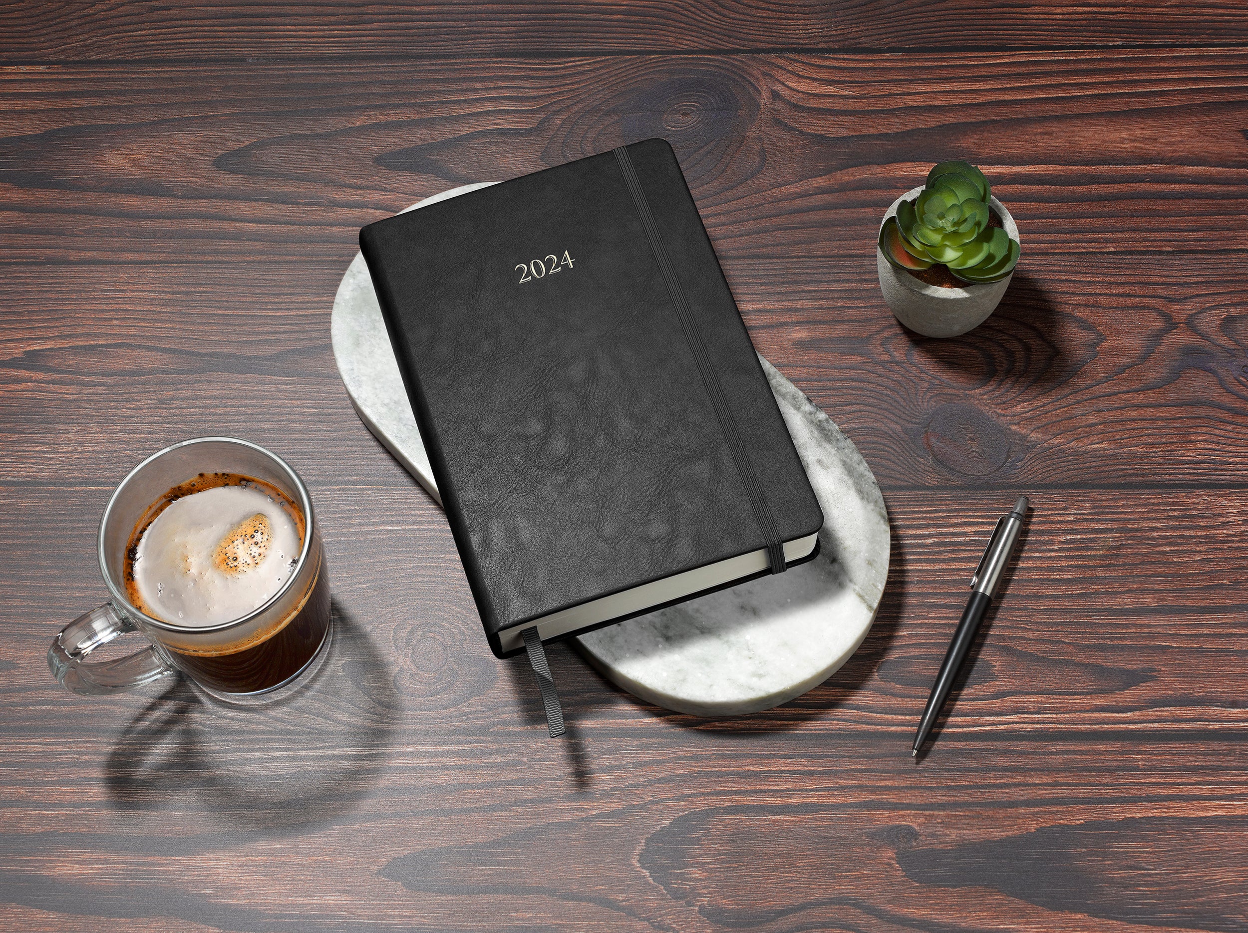 black 2024 daily journal in a5 sitting on desk with coffee and plant