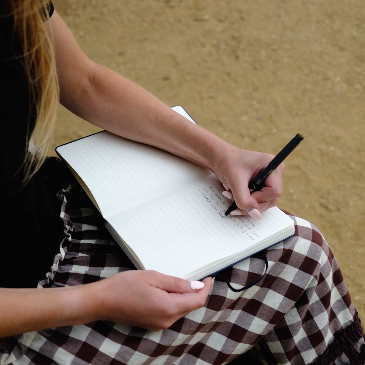 White-lined notebook open on a lap, with hands writing in the pages.
