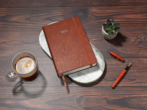brown 2024 daily journal in a5 sitting on desk with coffee and plant