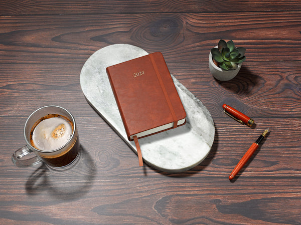 brown 2024 daily journal in a6 sitting on desk with coffee and plant