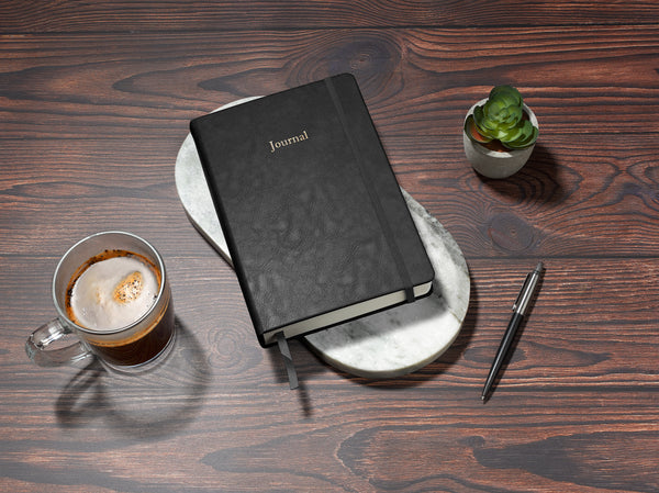 black undated daily journal in a5 sitting on desk with coffee and plant