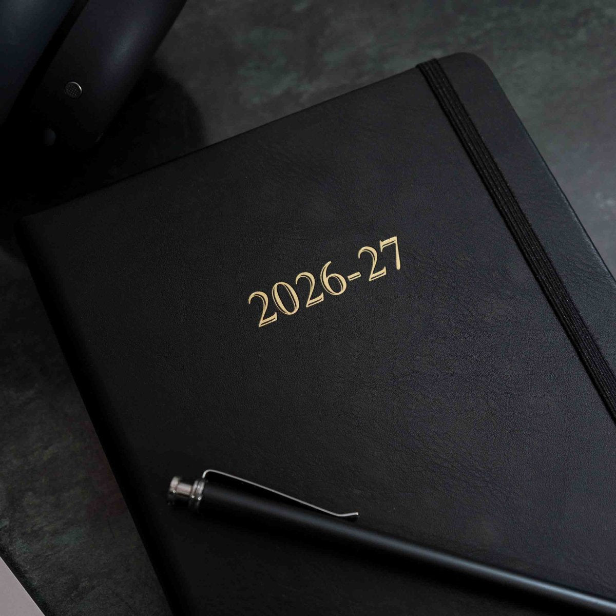Black executive journal with gold "2026-27" title and elastic strap, plus pen nearby